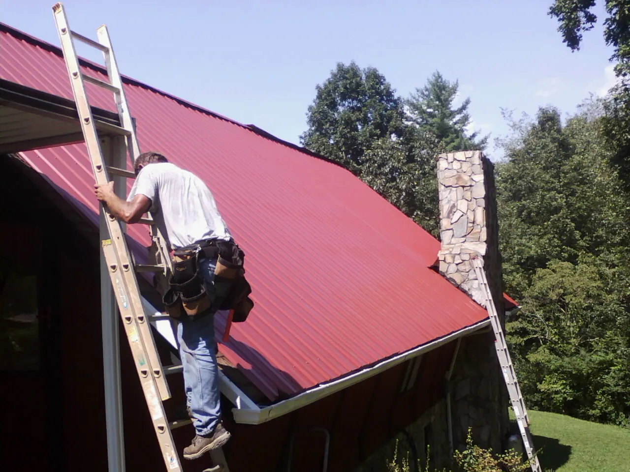 Roofer on a ladder beside a red metal roof and stone chimney — Soffit & Fascia Repair by Roofing Brothers in Gold Hill
